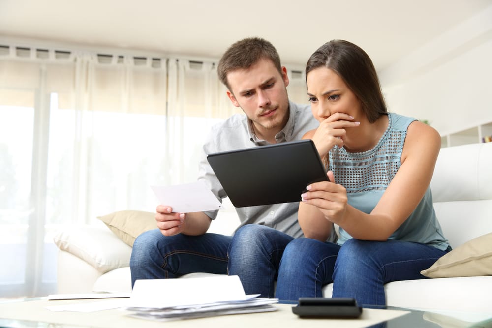 Worried couple checking bank account trouble online in a tablet to see if they can afford extras sitting on a couch in the living room at home
